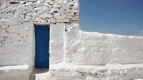 White stone wall with an old blue door. Background is bright blue sky. Street is visible (Mykonos, ...