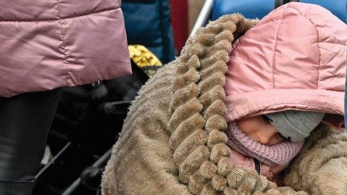 A child wrapped in a blanket sits on luggage while waiting to be relocated from the temporary ...