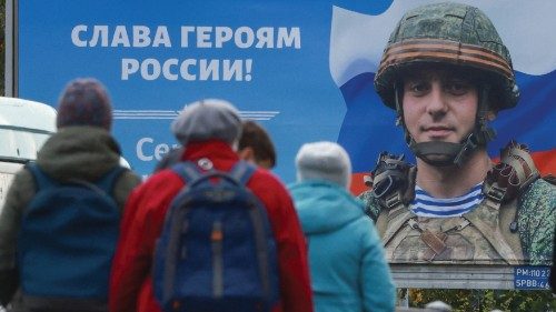 People gather at a tram stop in front of a board displaying a portrait of Russian service member ...