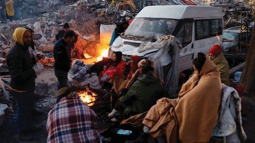 People sit around a fire next to rubble and damages near the site of a collapsed building in the ...
