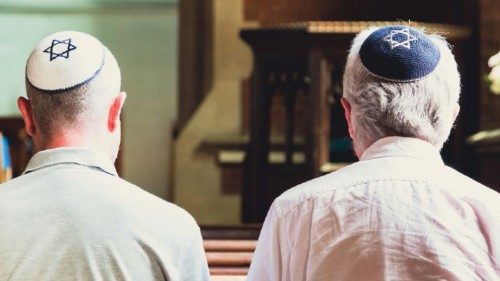 Close up image depicting a rear view of two Jewish men sitting together inside a synagogue. They ...